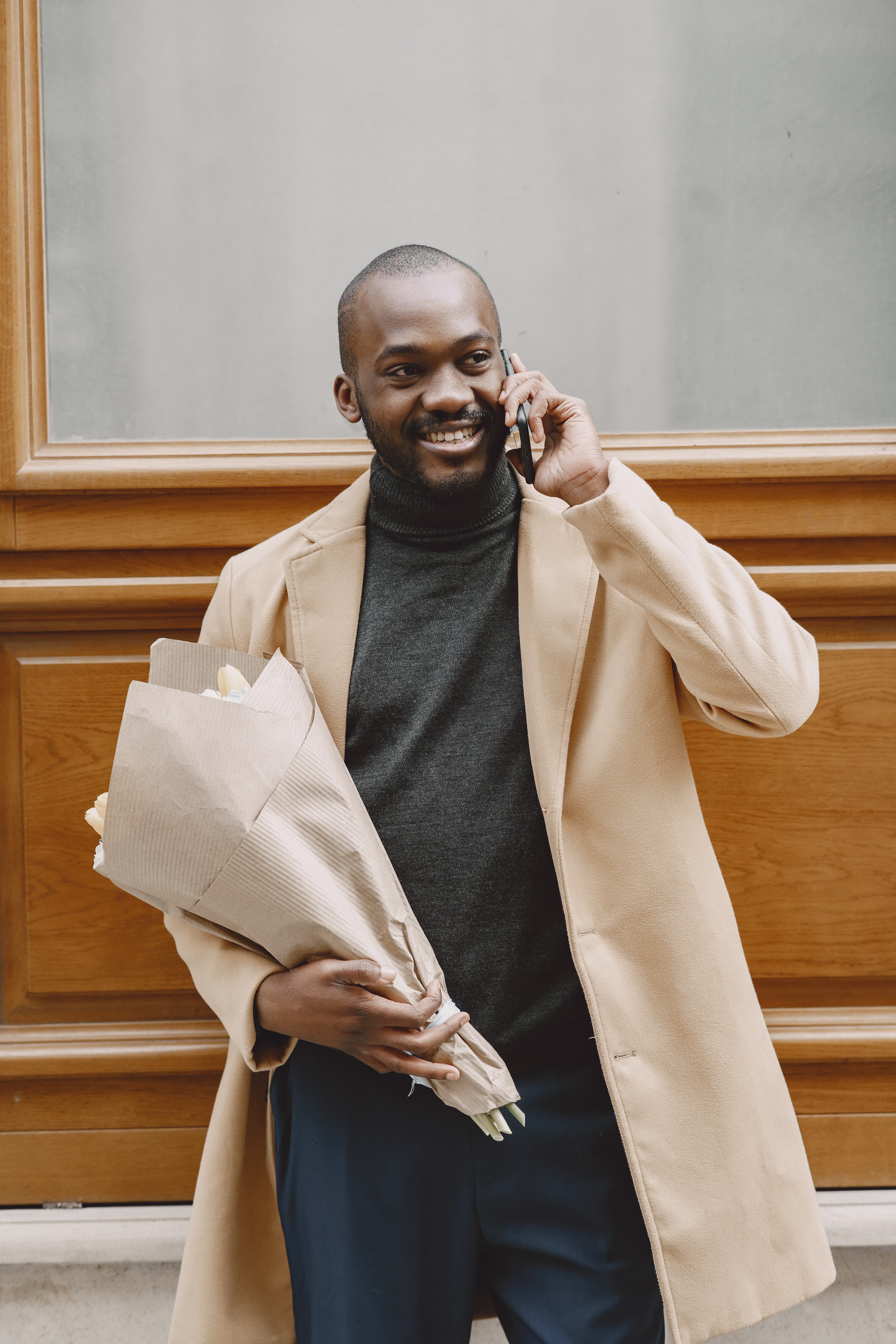 A smiling man in a camel coat on a phone call, holding a bouquet of flowers